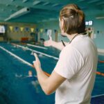 Male swimming coach standing by the swimming pool. He is showing staight hand to correct the student. Swimming technique, professional.